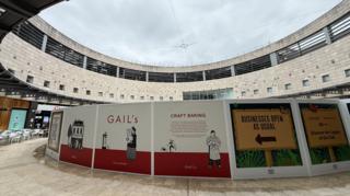 Hoarding for bakery chain Gail's erected in a circular courtyard. The hoarding has colourful branding on it, coloured white and red. It is cloudy above.