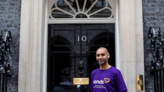 Man wearing purple Sands t shirt outside famous black number 10 door in Downing Street 