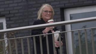 Sharman behind a metal silver fence walking into court. She is smiling and looking at the camera. She carries a takeaway coffee and a bag in her other hand.