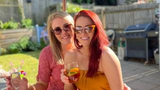 Friends Sarah (left) and Leanne (right) wearing sunglasses, smiling and holding drinks as they sit in a garden.