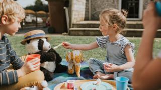 Children outside having a picnic with a teddy. A girl feeds the teddy some food on a spoon.