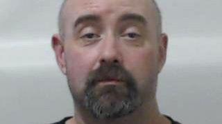 A police picture of a man with a bald head and brown beard against a grey background