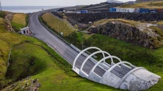 Aerial view of a modern road bridge with four white arch supports crossing a grassy ravine near a rocky coastline. The newly paved road curves toward the sea, with rugged cliffs, scattered buildings, and open water visible in the background.