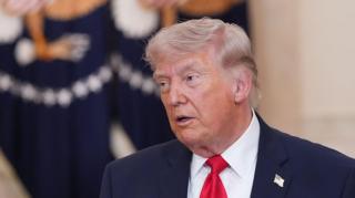 US President Donald Trump speaks while wearing a dark suit, white shirt and red tie. He stands indoors front of flag with a the US presidential seal on it.