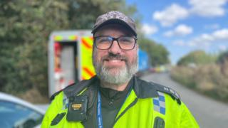 A man, Tom Carter, wearing a police uniform stood in a layby. He is standingin front of a van with hi-vis markings on it. He is smiling.