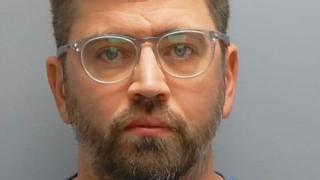 A mugshot of Della-Rocca who has fair hair, a grey brown beard, and grey-rimmed glasses.