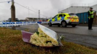 A bouquet of yellow roses placed on the ground with a police cordon in the background