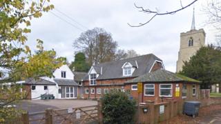 A primary school with a yard to the left and old two-storey brick buildings to the right. There is a white building with a triangular upper floor in the background, and a brick wall in the foreground with a wooden gate across the entrance. There is a church to the right.