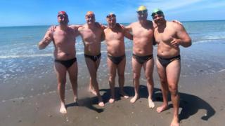 Five men wearing small black swimming pants, caps and goggles have their arms round each other and are smiling for the camera. They are standing on a beach in front of the sea.