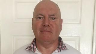 Philip Miller, who has a bald head and is wearing a white shirt with a dark red and black checked collar, is standing in front of a white door in a house. He looks impassive.