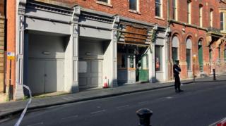 A police officer is standing on a road within a cordon outside a two-storey building. Police tape can be seen at one end of the cordon. Wooden doors to the building are open.