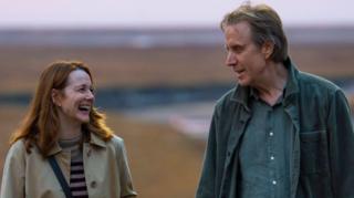 A screengrab of the film showing Rhys Ifans and Laura Linney looking at each other. Linney laughs while Ifans smiles at her. She wears a beige trench coat on top of a striped top, with her long brown hair reaching her shoulders. He wears a dark blue overshirt, with a blue shirt underneath and has short dark hair.