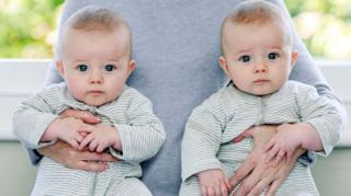 Two babies in green and white striped baby-grows. An adult is carrying them under either arm.
