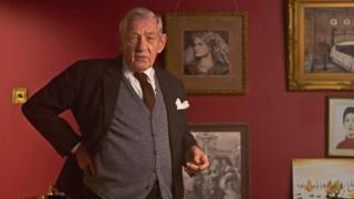 Ian McKellen stands in front of various paintings on a red wall, wearing a suit and waistcoat as he plays L.S. Lowry.