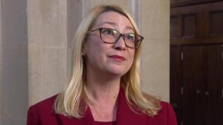 Jess Asato during an interview. She looks to the right of the camera. She has shoulder length blonde hair, glasses and is wearing a red blazer.