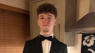 A teenage boy stood in a formal suit, including a black jacket and black bow tie, stood in a kitchen, smiling for the camera.