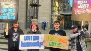 Four people, three men and a woman, hold signs outside a building. Their signs read "unions ain't busted" and "union busted?" and "unions protect workers" and "grand theft wages solidarity"