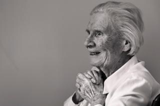 A black and white photo of an elderly lady sitting in a chair against a plain wall, smiling with her hands clasped as she looks out of the frame.