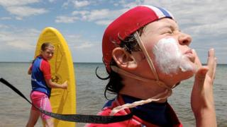 A young child in a swimming cap applying sunscreen on their face, with another young child holding a surfboard, with the ocean in the background.