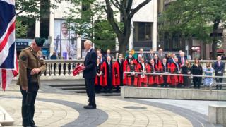 To the left there is a union flag.
An armed forces representative dressed in a brown military uniform bowing his head.
In the centre of the photo is a man with a dark suit and grey hair, who is holding a poppy wreath.
In the background there are people dressed in red gowns lined up watching the ceremony.