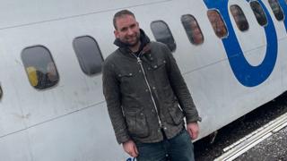 A man wearing a brown jacket and jeans standing in front of a white plane fuselage.