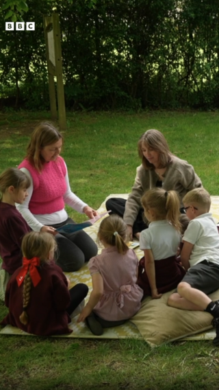 A group of children and two adults sitting on a blanket in a green space, surrounded by trees.