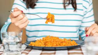 A person sitting at a table eating penne pasta from a plate with a fork.