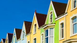 A row of closely spaced terraced houses viewed from a low angle, emphasizing their sloping roofs and upper floors, on a bright sunny day with a blue sky.
The houses are painted in bright, contrasting pastel colours - including light blue, pale yellow and a vivid lime green.
