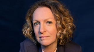 A professional headshot of Victoria Woodall with chin length golden curly hair wearing a navy suit against a navy backdrop