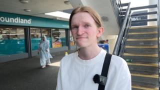 A young white man with reddish blonde hair tied back smiles at the camera. He is in the street, and wears a white top. A branch of Poundland is pictured behind him, as is a concrete flight of stairs.