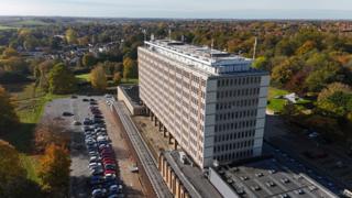 A drone image of a large office building in a rectangular shape, with trees, a car park and housing around it.
