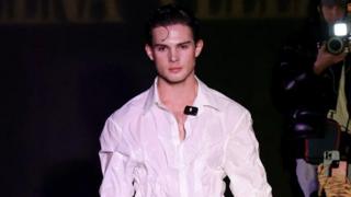 Braden Peters stares straight ahead as he walks a runway in a white button-up shirt.
