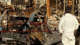 A person in a protective suit walks through the aftermath of the fires in LA. A car surrounded by debris and charred remains of buildings