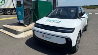 The Shell Triple 10 Challenge Concept Car is white with black wheels, window panes and rims. It is parked in front of an electric vehicle charger on a sunny day.