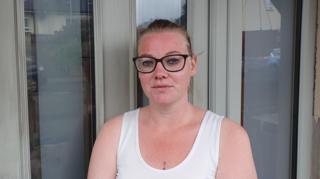 Roberta, a woman with light brown hair tied back from her face, is looking at the camera. She is wearing a white vest top and black framed glasses.