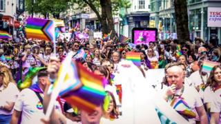 People marching in the 2024 Birmingham Pride parade, through the city centre, Many are carrying rainbow flags and some are blowing whistles.