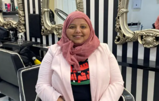 Shaista is pictured in a hairdresser's chair, smiling at the camera, with mirrors behind her. She has a dark pink headscarf and is wearing a light pink jacket 