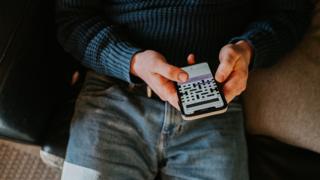 A man, wearing blue jeans and a dark blue shirt, at home holds a smart phone and uses a cryptic crossword app.