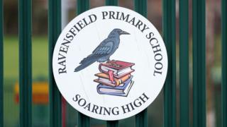 A close-up view of the Ravensfield Primary School sign on dark green railings. The sign is circular and features a drawn picture of a raven bird sitting on a pile of three books on a white background. Around the edge of the circle are the words "Ravensfield Primary School" at the top and "Soaring high' at the bottom.