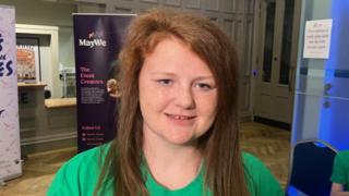 Jamiee-Leigh is smiling at the camera and has long ginger hair. She is wearing a bright green top.