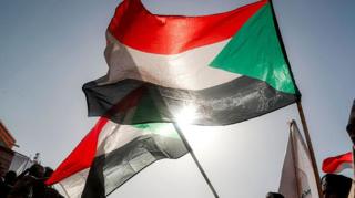 A Sudanese flag flutters in the wind against a backdrop of a bright blue sky - other Sudanese flags are in the background. The sun can be seen shining through the bottom of the flag.