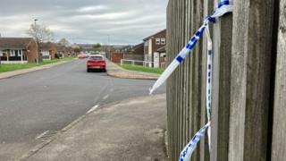A residential street with a fence with blue and white police tape tied around it on the right hand side. Homes on either side of the street and cars parked along the street in the distance.
