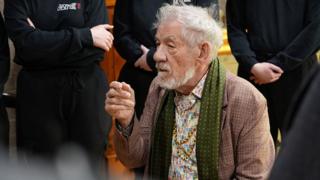 Sir Ian McKellen is an 86-year-old man wearing a tweed jacket, a green silk scarf and a flowery shirt with two long gold necklaces. He is seated and behind him are a number of people wearing black who are standing listening although only their torsos are in shot 