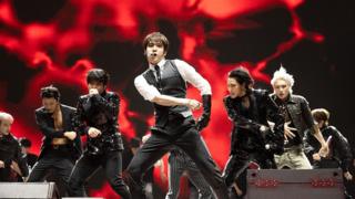 Members of Ateez perform onstage. Seven of the eight members are visible, with the lead in front wearing a white shirt, black glove and grey and black waistcoat. The background is red and black.
