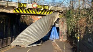 A damaged railway bridge with a piece of metal hanging down onto the road below and the words "low bridge" in large yellow lettering
