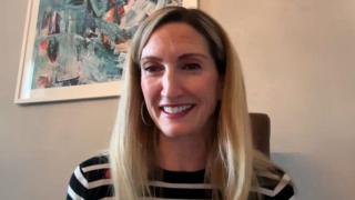 Jjoy Dunlop, Stiùiriche Sheachdain na Gàidhlig agus i na suidhe air beulaibh camera agus lèine dubh is geal oirre, falt fada bàn agus dealbh air a' bhalla air a cùlaibh.