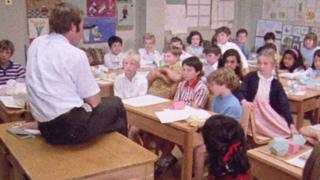 A classroom of children sit around a man on a teacher's desk.
