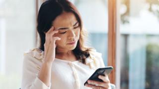 A woman, looking down stressed at her phone while at home. She is wearing a cream coloured top.