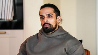 A still from EastEnders showing the character of Ravi, wearing a grey hoodie and looking into the distance.