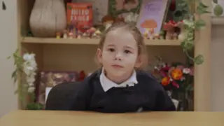 A young girl is in school uniform at a desk looking at the camera